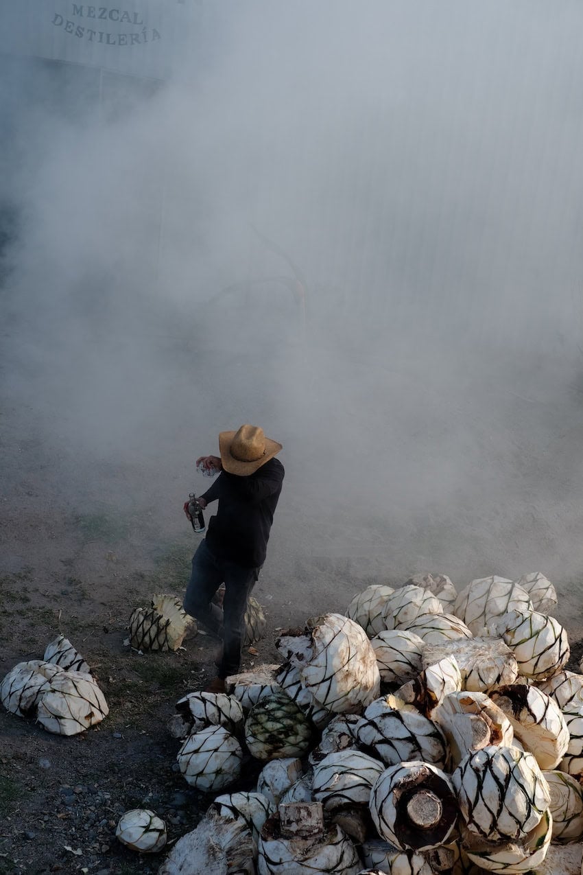 Smoke goes into the eyes of a worker at the mezcal palenque while he holds onto a bottle