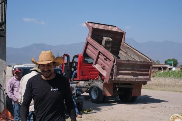 A man smiling in the foreground of a photo of a tip truck that has delivered agave plants, imaginably to the factory.