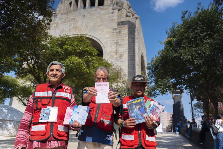 A group of 3 men wearing red vests selling a street paper in Mexico City.