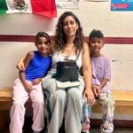 A mother and her two children sitting on a bench beneath a Mexican flag, waiting for documentation from Soy México