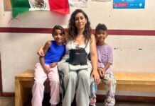A mother and her two children sitting on a bench beneath a Mexican flag, waiting for documentation from Soy México
