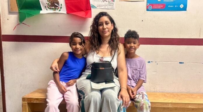 A mother and her two children sitting on a bench beneath a Mexican flag, waiting for documentation from Soy México
