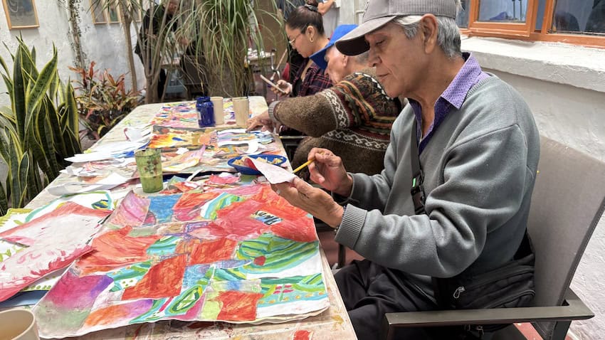 An elderly man drawing on a piece of paper with a larger art piece on the table in front of him.