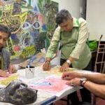 A group of 3 men sitting around a table creating artwork with a colorful mural in the background.
