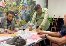 A group of 3 men sitting around a table creating artwork with a colorful mural in the background.