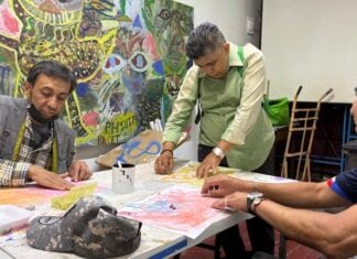 A group of 3 men sitting around a table creating artwork with a colorful mural in the background.