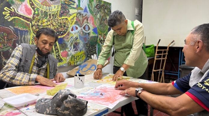 A group of 3 men sitting around a table creating artwork with a colorful mural in the background.