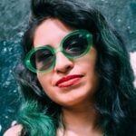 Young Hispanic woman with green hair tints and wearing green glasses.