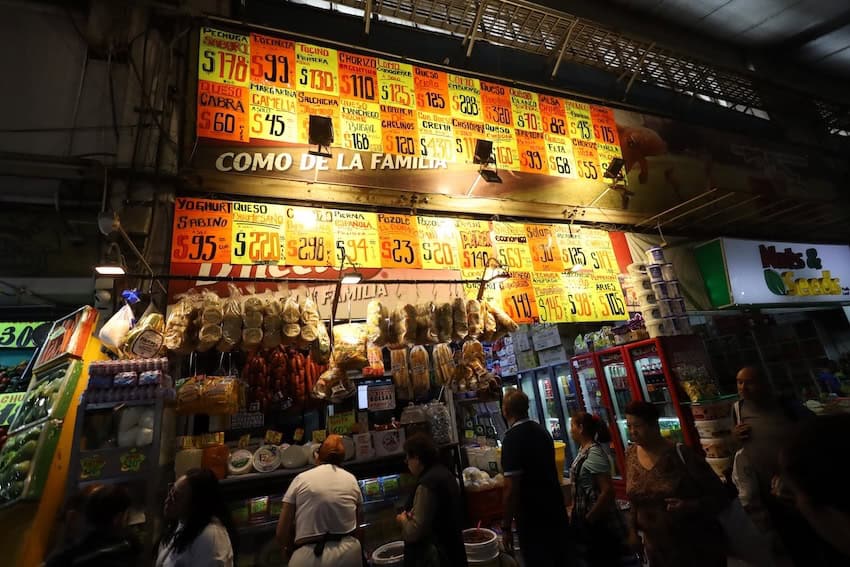 A small market stall lists prices of each item on a large board above food stuffs. People are waiting in line for service below.