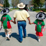 A family celebrates Cinco de Mayo in Port Huron, Michigan. A man in a cowboy hat holds the hands of two young boys in mariachi sombreros, holding US flags