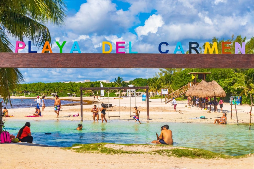 Tourists lounge in a beachside pool under a sign reading Playa del Carmen